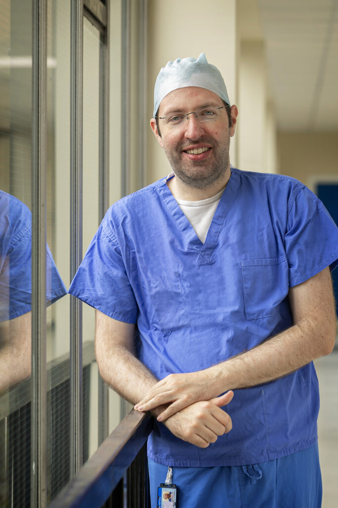 Image of Dr Matthew Costley stood in blue scrubs with a surgical hat on in a white corridor near windows. 