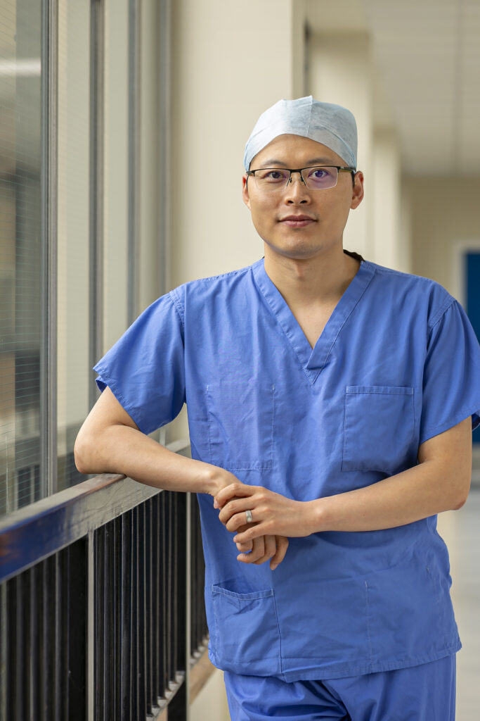 Picture of Dr Shaochen Liu in blue scrubs and hair net with elbow leant on a railing. 