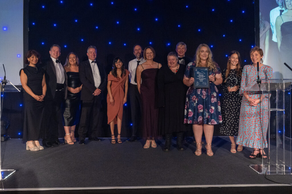 Picture of the BadgerNet Maternity Support Team stood on stage posing for a photo holding the award and certificate in front of star backdrop, with comedian Shaparak Khorsandi, Chairman Professor Sir John Burn and Maurya Cushlow.