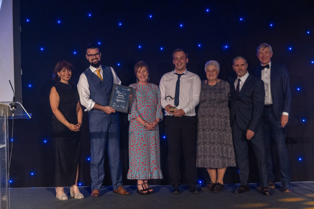 Picture of the Simulation Team stood on stage posing for a photo holding the award and certificate in front of star backdrop, with comedian Shaparak Khorsandi, Chairman Professor Sir John Burn and Maurya Cushlow.