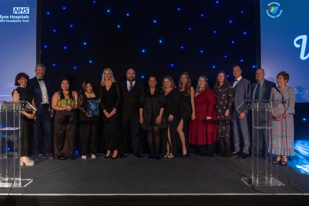 Picture of the International Recruitment Steering Group stood on stage posing for a photo holding the award and certificate in front of star backdrop, with comedian Shaparak Khorsandi, Chairman Professor Sir John Burn and Maurya Cushlow.