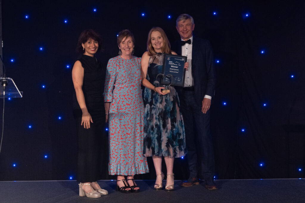 Picture of Sinead Greener stood on stage posing for a photo holding the award and certificate in front of star backdrop, with comedian Shaparak Khorsandi, Chairman Professor Sir John Burn and Maurya Cushlow.
