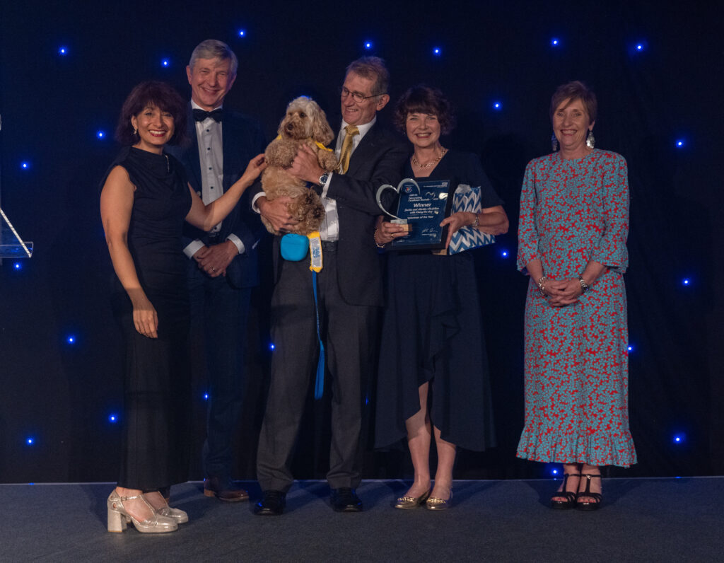 Picture of the Jackie and Martin McLellan and Daisy the dog stood on stage posing for a photo holding the award and certificate in front of star backdrop, with comedian Shaparak Khorsandi, Chairman Professor Sir John Burn and Maurya Cushlow.