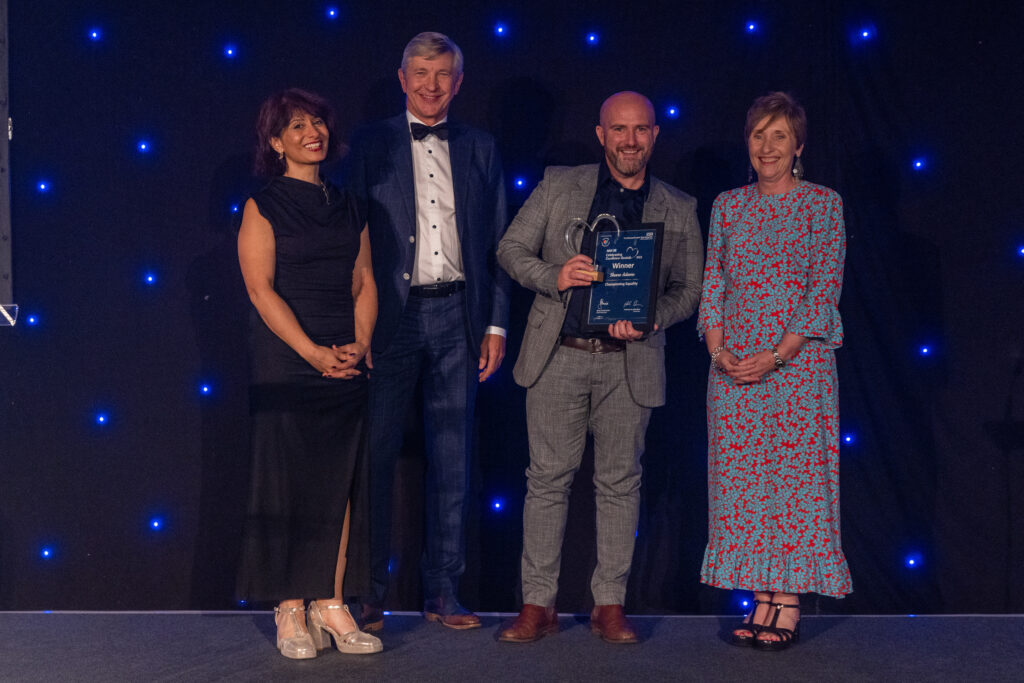 Picture of the Shane Adams stood on stage posing for a photo holding the award and certificate in front of star backdrop, with comedian Shaparak Khorsandi, Chairman Professor Sir John Burn and Maurya Cushlow.