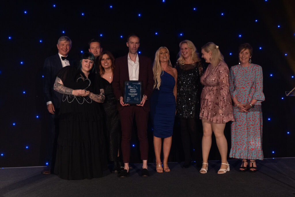 Picture of the Procurement and Supply Chain team stood on stage posing for a photo holding the award and certificate in front of star backdrop with Chairman Professor Sir John Burn and Maurya Cushlow.