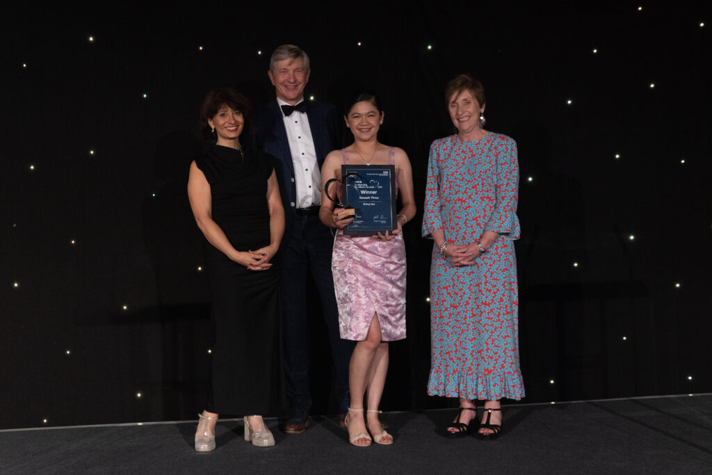 Picture of the Hannah Perez stood on stage posing for a photo holding the award and certificate in front of star backdrop, with comedian Shaparak Khorsandi, Chairman Professor Sir John Burn and Maurya Cushlow.