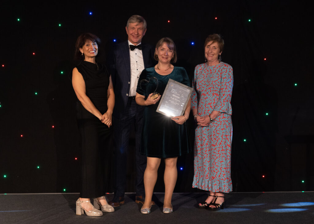Picture of the Paula Newman stood on stage posing for a photo holding the award and certificate in front of star backdrop, with comedian Shaparak Khorsandi, Chairman Professor Sir John Burn and Maurya Cushlow.