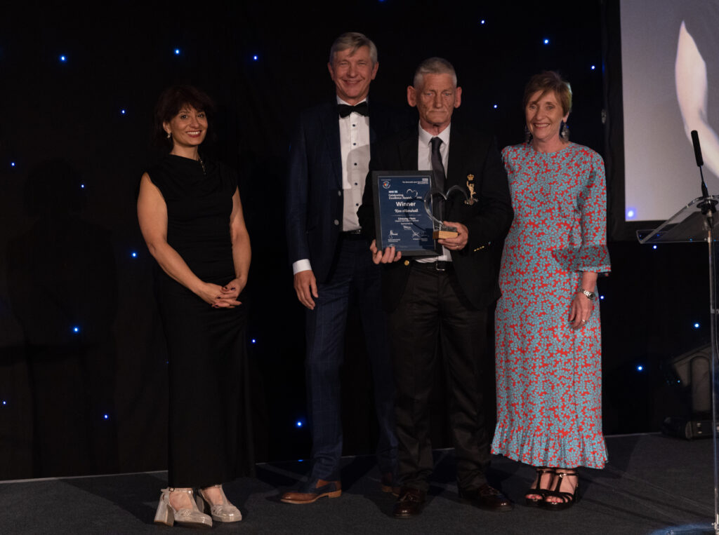 Picture of the Ken Marshall stood on stage posing for a photo holding the award and certificate in front of star backdrop, with comedian Shaparak Khorsandi, Chairman Professor Sir John Burn and Maurya Cushlow.