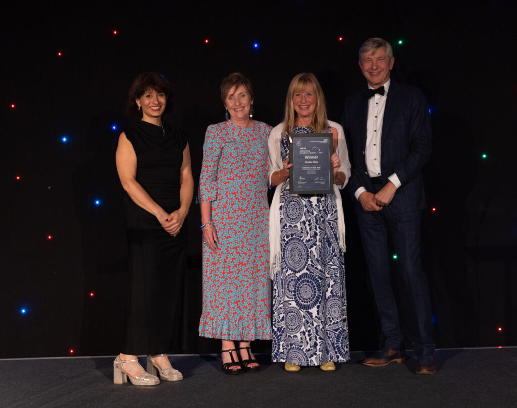 Picture of Jackie Rees stood on stage posing for a photo holding the award and certificate in front of star backdrop, with comedian Shaparak Khorsandi, Chairman Professor Sir John Burn and Maurya Cushlow.