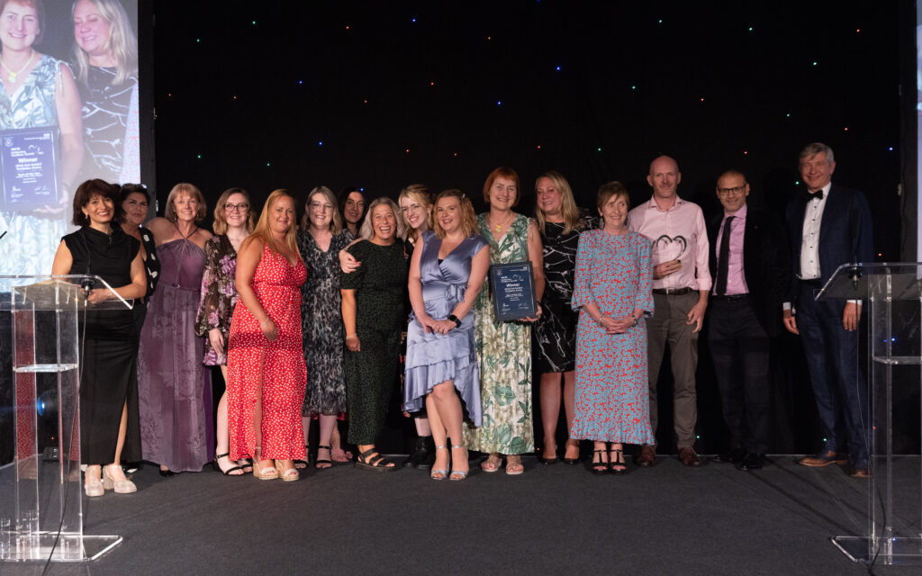 Picture of the North East Assisted Ventilation Service on stage posing for a photo holding the award and certificate in front of star backdrop, with comedian Shaparak Khorsandi, Chairman Professor Sir John Burn and Maurya Cushlow.