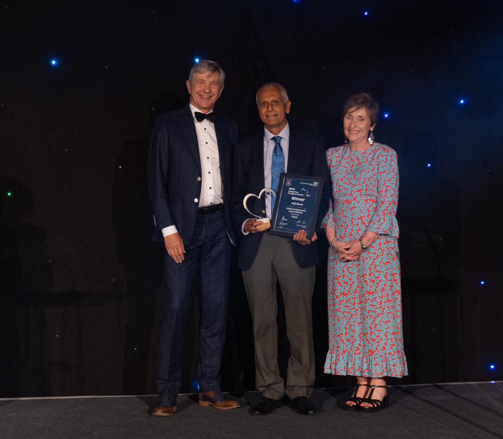 Picture of Asif Hasan stood on stage posing for a photo holding the award and certificate in front of star backdrop, with Chairman Professor Sir John Burn and Maurya Cushlow.