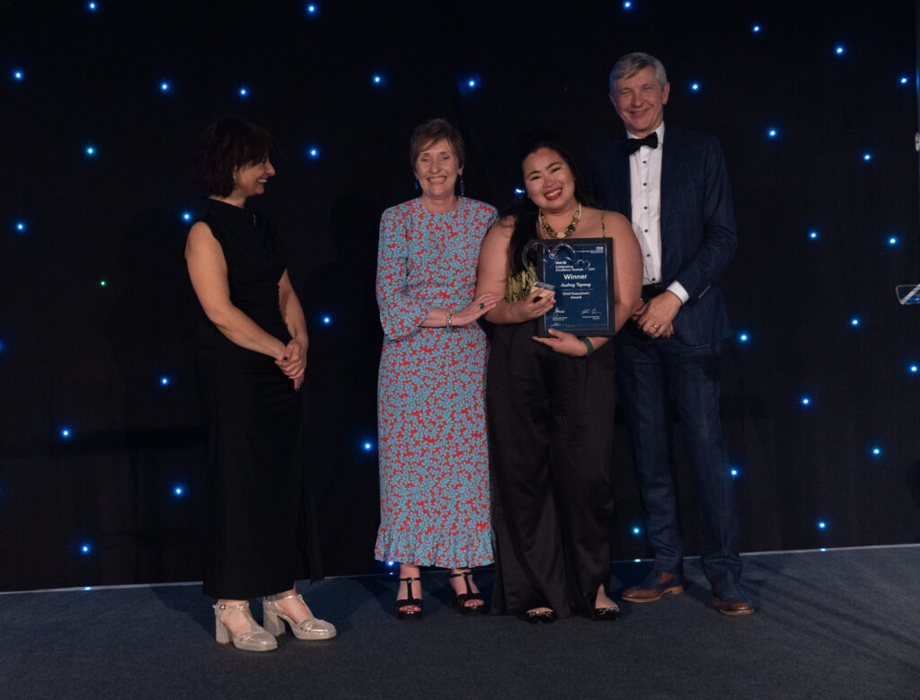 Picture of Audrey Tapang stood on stage posing for a photo holding the award and certificate in front of star backdrop, with comedian Shaparak Khorsandi looking at her, and Chairman Professor Sir John Burn and Maurya Cushlow.