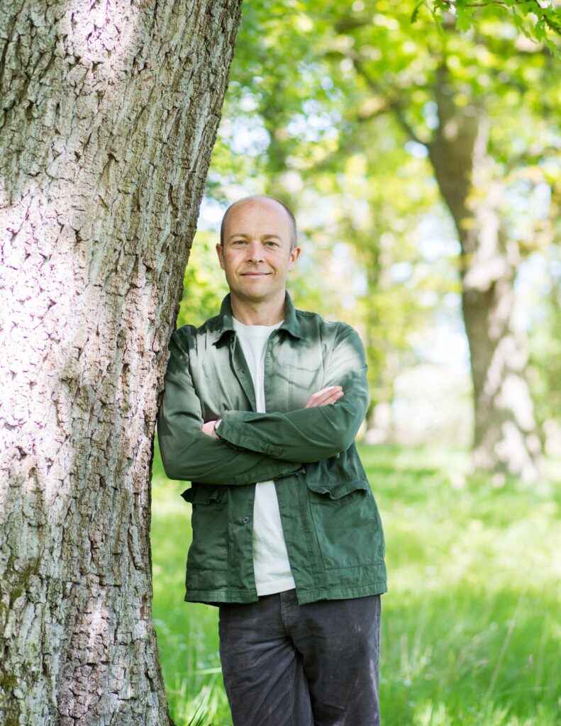 Nick James is outdoors and leans against a tree with his arms crossed.