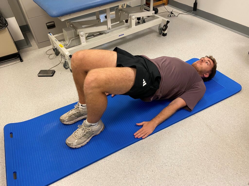 Picture of someone lying on a blue mat on their back with your knees bent and feet flat on the floor and pushing their buttocks off the floor.