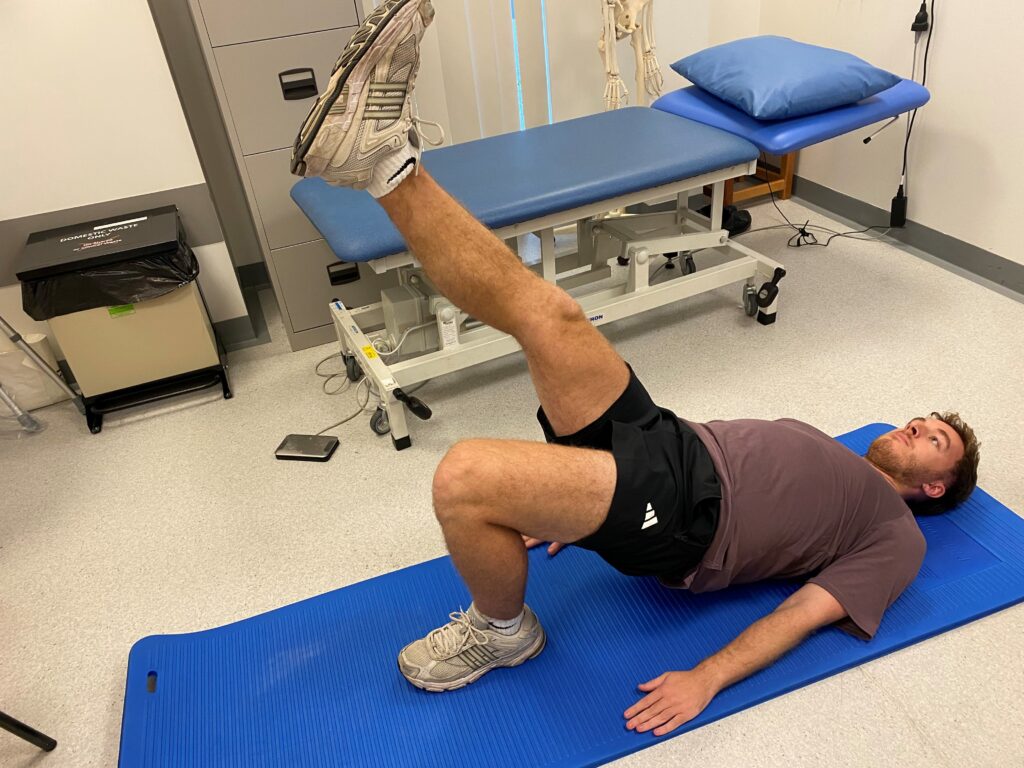 Picture of someone lying on a blue mat on their back with one knee bent and the other leg extended pushing up your buttock off the floor.