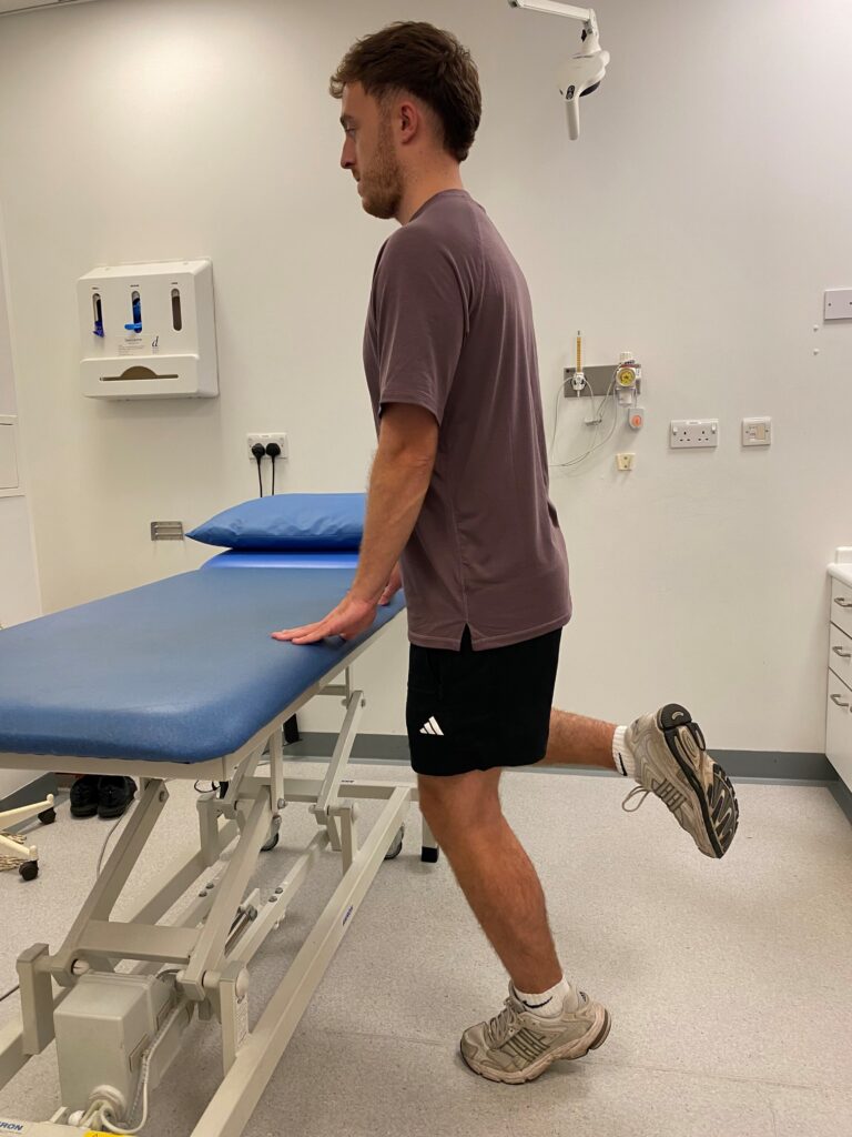 Person stood on one leg, with one leg bent holding onto a bed.