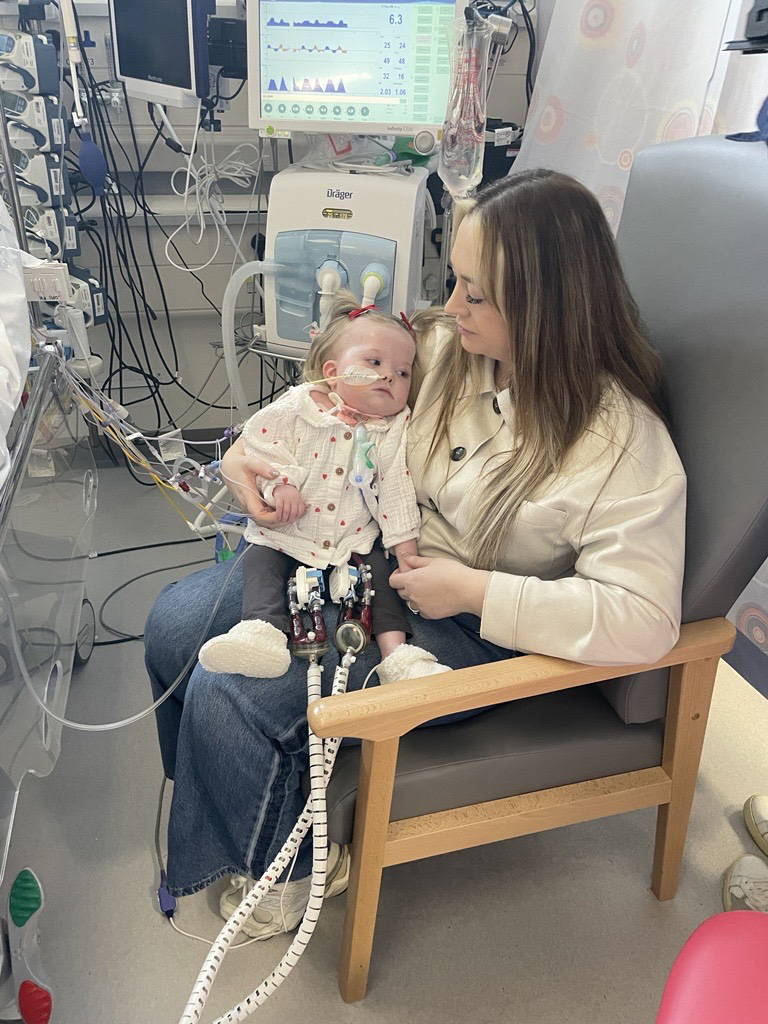 Picture of Poppy with biventricular assist device sat on her mum, Becca's, knee in a hospital chair in the Freeman Hospital. 