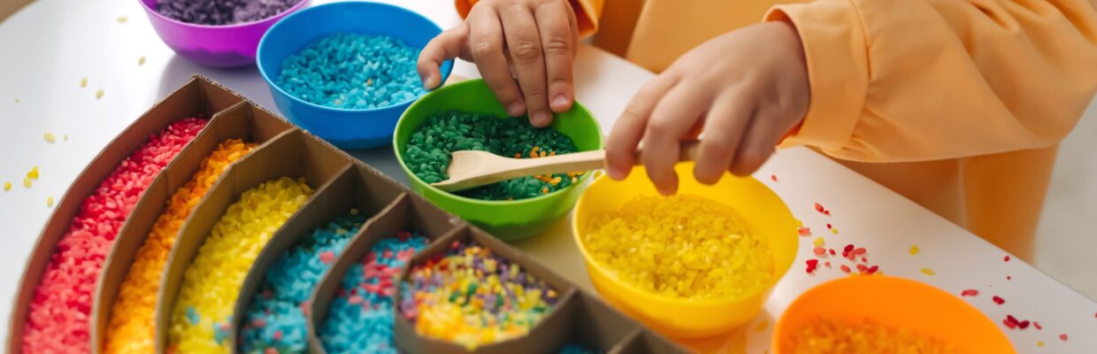 Child playing with coloured rice in different coloured bowls, putting them into a rainbow shape.