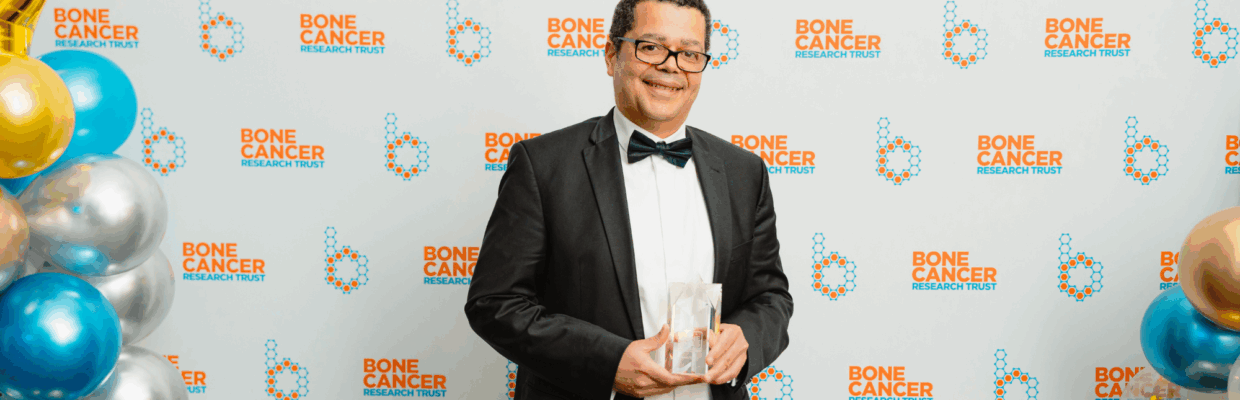Mr Kenny Rankin stands against a white background with the Bone Cancer Research Trust. He is holding his award.