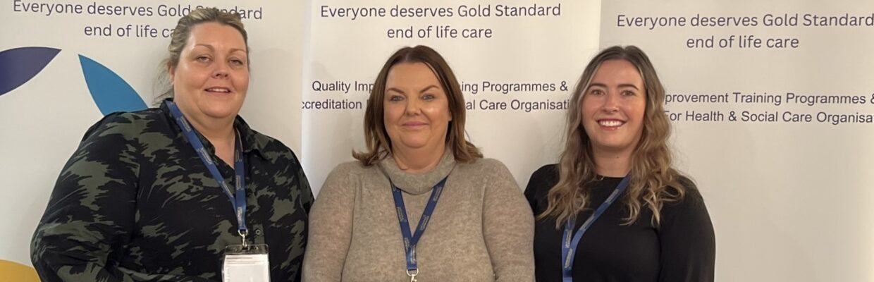 Three women standing together in front of a banner with the text "Everyone deserves Gold Standard end of life care"