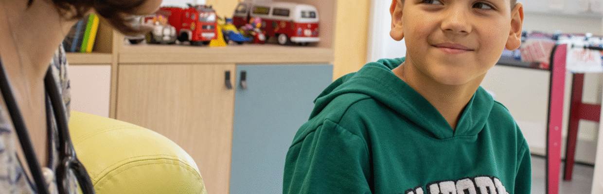 Picture of a young boy sat down looking to the left at a member of staff in a clinical setting. There are toys on a shelf in the background of the shot.