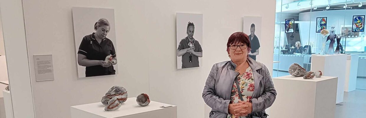 Picture of Anne Scott stood in front of glass artwork on white platform, with photos of NHS staff hung on the wall.