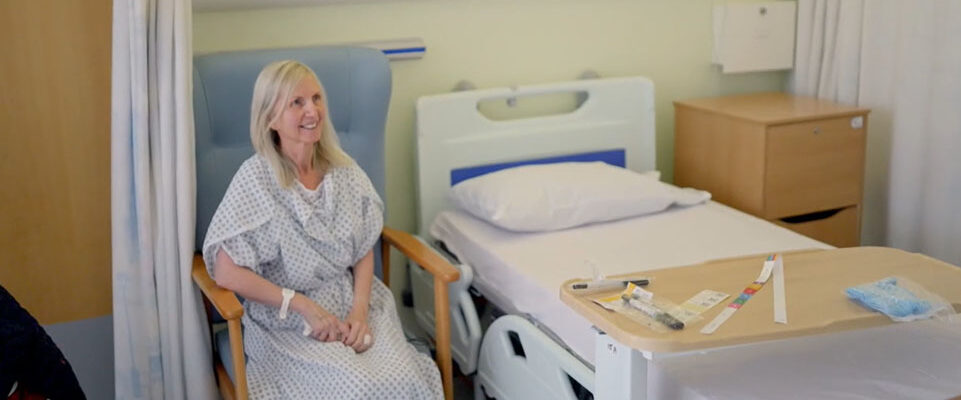 Picture of Susan sat on blue chair in surgical gown on the ward.