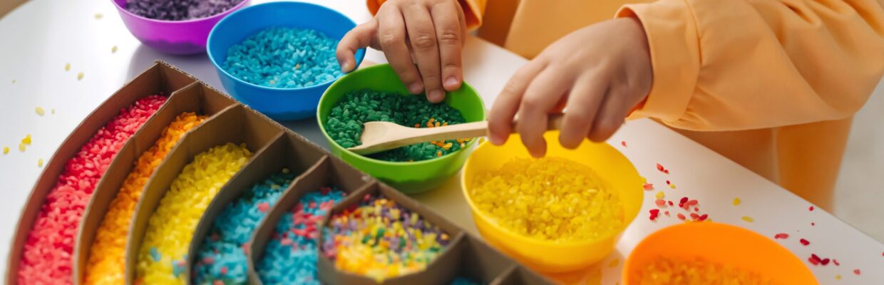 Child playing with coloured rice in different coloured bowls, putting them into a rainbow shape.