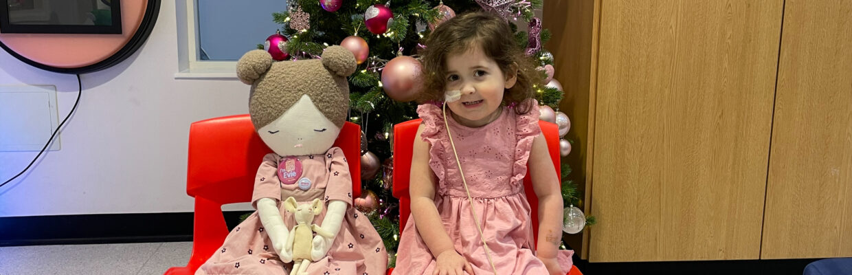Image shows 4-year-old Evie sitting on a chair next to her doll. They are in front of a Christmas tree.
