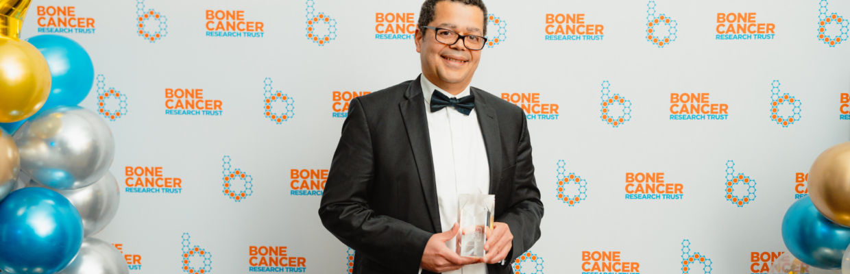 Mr Kenny Rankin stands against a white background with the Bone Cancer Research Trust. He is holding his award.